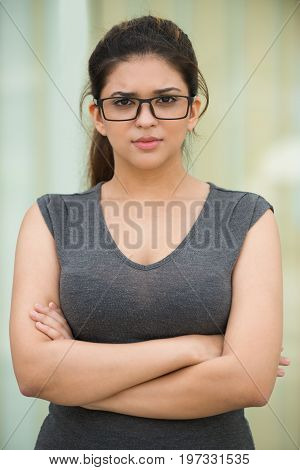Portrait of serious young Caucasian businesswoman or student wearing grey dress and glasses standing with crossed arms and looking at camera outdoors