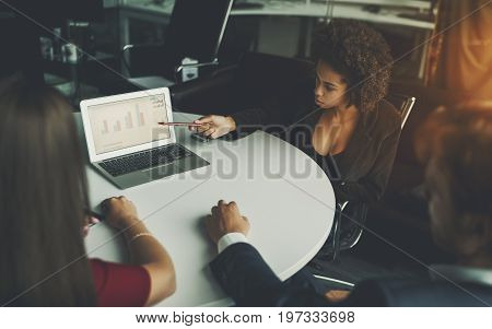 Young cute afro american woman probationer in meeting room is showing graph of her business project development to her boss and colleagues on screen of laptop during work meeting at oval table