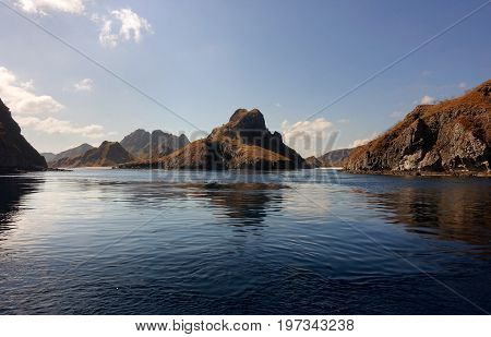 Coastline Of Mountains With Green Vegetation Reflected  In Blue Ocean Water At Labuan Bajo In Flores