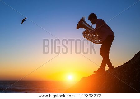 Silhouette of a young musician playing the Tuba on rocky sea coast during sunset.