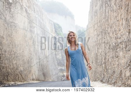 Attractive smiling girl in light-blue dress is standing on the road between mountains and looking in the camera