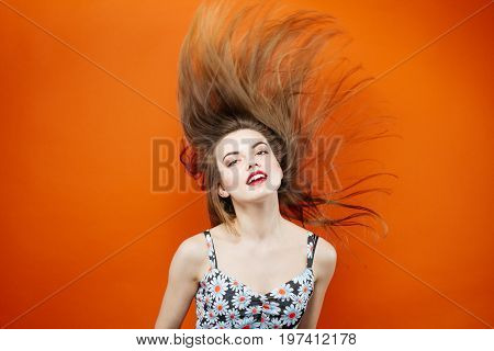Temperamental Woman With Long Dark Hair Wearing Dress With Flowers Is Having Fun In Studio On Orange