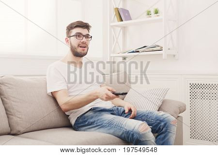 Young man wathing tv at home. Amused guy pointing with remote controller on tv-set, copy space