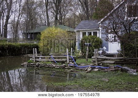 Close up landscape view of flood waters rising over a paved road in front of properties with trees foliage and the houses partly submerged in the distance in the West Island of Montreal, Quebec on a bright overcast day in May.