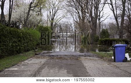 Landscape view of flood waters rising over a paved road in front of a gated property with trees foliage and a house partly submerged in the distance in the West Island of Montreal, Quebec on a bright overcast day in May.