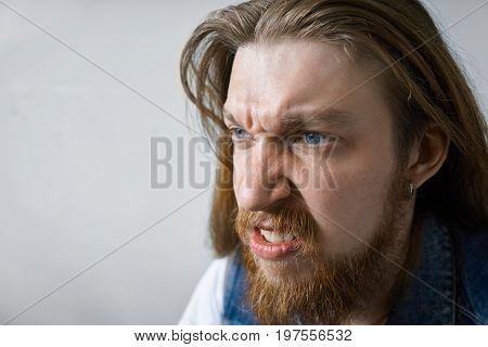 Close up studio portrait of angry mad young man with thick beard and long hairstyle frowning and clenching teeth his eyes full of anger and fury. Negative human emotions aggression rage and frenzy