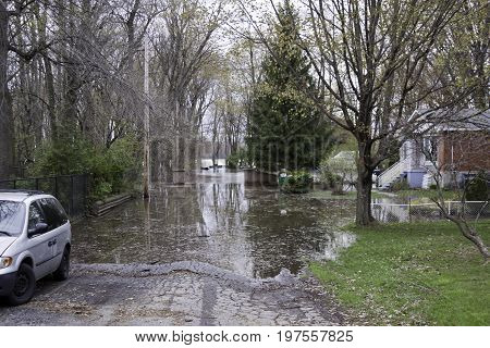 Landscape view of flood waters rising over a paved road with trees foliage and a house partly submerged in the West Island of Montreal, Quebec on a bright overcast day in May.