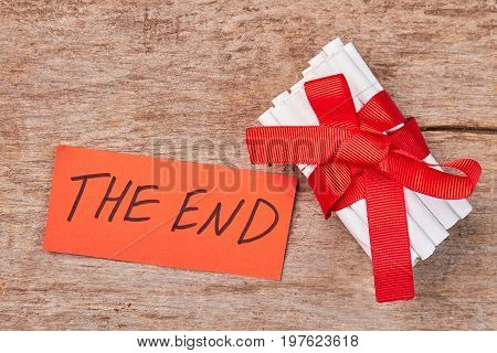 Message the end, tobacco cigarettes. Pile of white toxic cigarettes with red bow and ribbon, wooden background.