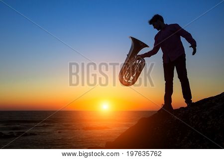 Silhouette of musician with the trumpet on rocky sea coast during amazing sunset. Tuba instrument. 