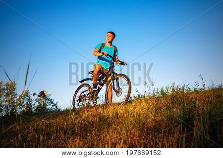 The boy on a bicycle is looking the sunset. The boy is standing on the hillside, behind him a blue sky without clouds.