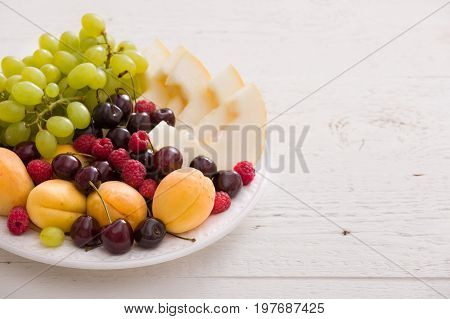 Assortment of juicy fruits on white plate and white table background. Organic raspberries apricots melon cherries - summer dessert or snack. healthy eating concept.