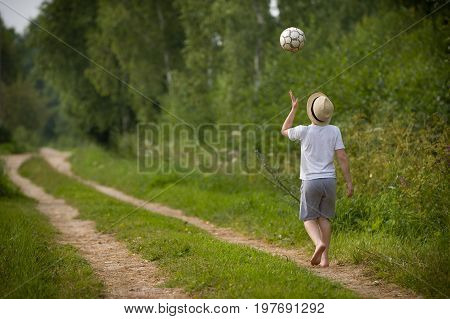 Back view on kid boy in a straw hat walking on a countryside road and playing with football ball. Child outdoors on a sunny summer day. Lifestyle vacations concept.