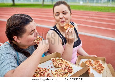 Happy young over-size women eating tasty and appetizing pizza after workout on stadium