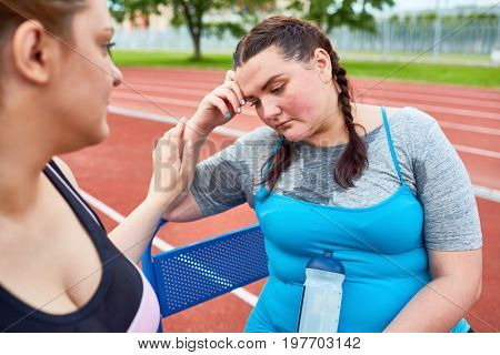 Sad woman with bottle of water sitting on bench after tiresome workout while friend encouraging her not to quit training