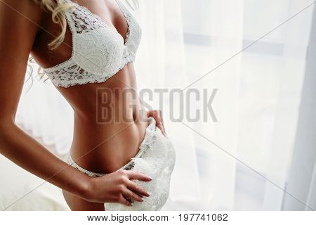 Sexual bride standing near the window in white lace lingerie is putting on her white wedding dress in luxury room. Close-up. Morning preparetion