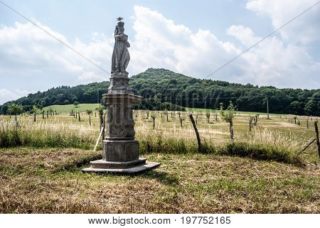 statue of Sv. Antonin Paduansky built in 18th century near Kocourov village in Ceske Stredohor mountains in Czech republici with young orchard andLhota hill on the background during nice summer day with blue sky and clouds