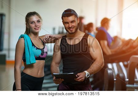 Happy smiling young woman standing with personal trainer in gym. Male coach and female client looking at the camera and posing on the background of treadmills. Fitness and healthy lifesyle concept.