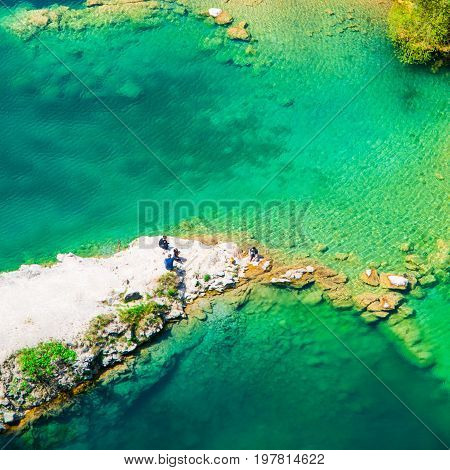 Rocky ness in blue azure water ang group of people having a picnic on it.