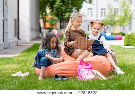 adorable multiethnic schoolgirls sitting on bean bag chair and reading book on schoolyard