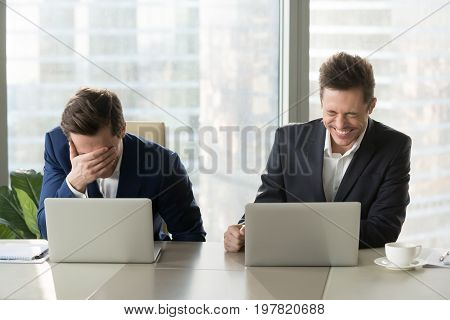 Two businessmen laughing out loud at workplace, office workers screaming with laughter and can not stop, funny positive emotions at work, cheerful colleagues having fun sitting at desk with laptops