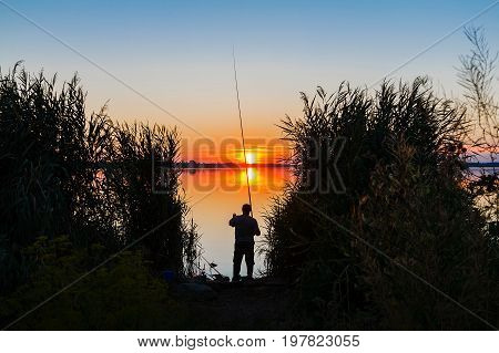 Sunset over the lake. On the shore is a fisherman and casts a fishing pole.