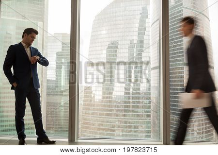 Businessman waiting for unpunctual partner being late for meeting, standing in office with financial district city view at background, looking at wristwatch, checking time while someone hurrying up