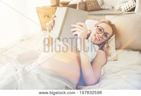 Time for rest. Cheerful pregnant young woman lying in bed and smiling while reading book