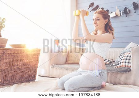 Healthy way of life. Cheerful glad pregnant woman smiling and holding dumbbels while doing sport exercises