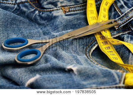 Tailors tools on denim textile. Metal scissors and yellow measure tape on jeans. Jeans belt loops zipper and pocket close up selective focus. Making clothes and design concept.