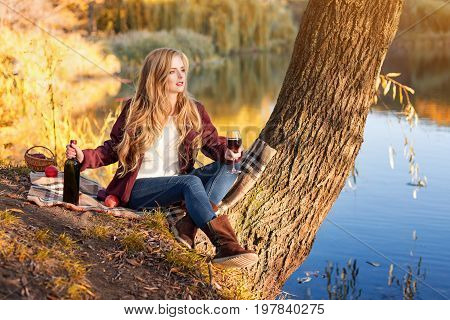 Beautiful woman drinking wine outdoors having picnic in the park. Portrait of young blonde beauty enjoying a glass of red wine sittind on autumn lawn near the lake