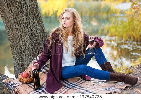 Beautiful woman drinking wine outdoors having picnic in the park. Portrait of young blonde beauty enjoying a glass of red wine sittind on autumn lawn near the lake