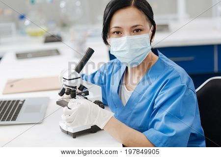 Working in the lab. Pleasant nice professional biologist wearing uniform and sitting at the table with a microscope while working in the lab