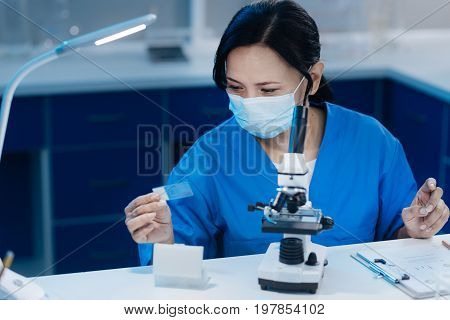Biological testing. Intelligent smart female biologist sitting at the table and using microscope while studying test sample slides