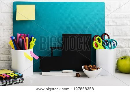 School background: a white brick wall with accessories, pencils, tetrads, scissors, an apple, pecans, a board for a substitute placed in front of her on the table. Selective focus