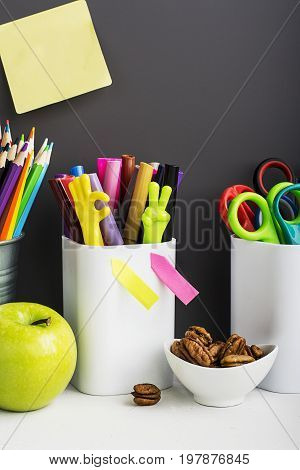 School background: a white brick wall with accessories, pencils, tetrads, scissors, an apple, pecans, a board for a substitute placed in front of her on the table. Selective focus