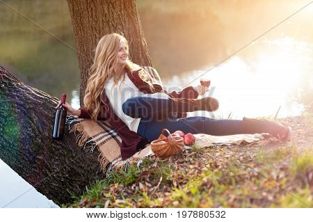 Beautiful woman drinking wine outdoors having picnic in the park. Portrait of young blonde beauty enjoying a glass of red wine sittind on autumn lawn near the lake