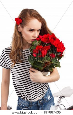 Young unemotional woman in casual clothing smelling flowers in pot and leaning on bicycle.