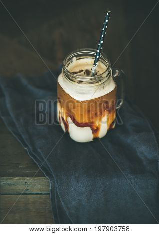 Iced caramel macciato coffee with milk in glass jar over dark rustic wooden table background, selective focus, copy space. Cold sweet refreshing summer drink concept
