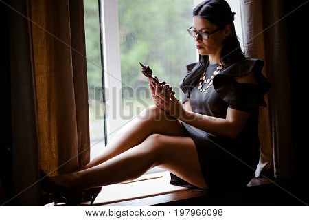 Brunette woman sitting on window sill. Female relaxing, thinking holding statuette of ioriental diety in her hands. Toned image.