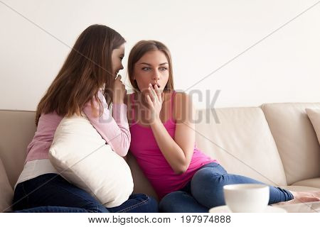 Two close girlfriends sharing scandalous news. Young attractive woman telling incredible secret to her friends ear while sitting on sofa in living room. Surprised lady listening interesting gossips
