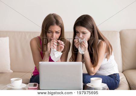 Two sentimental women friends upset crying and wiping tears with handkerchiefs while watching dramatic, sad movie, TV reality show or touching old home video on laptop while sitting on sofa at home