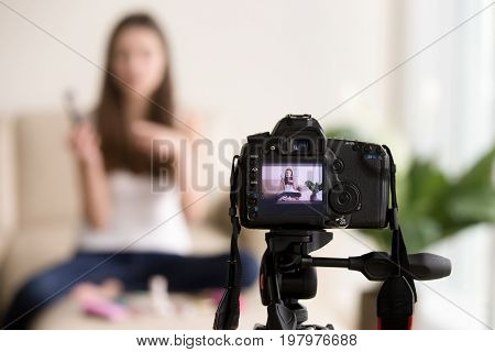 Close up photo of digital camera on tripod with young woman using cosmetics image on LCD back screen and blurred scene on background. Female video blogger records vlog, beauty vloger streaming online