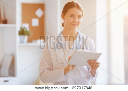 Waist Up Portrait Of Young Female Doctor Looking At Camera While Posing In Office Lit By Warm Sunlig