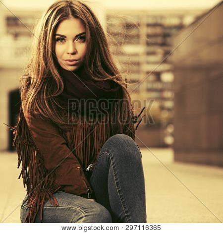 Young fashion woman sitting on city street sidewalk  Stylish female model in brown leather fringe suede jacket and scarf