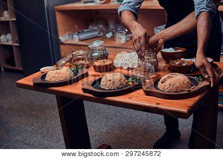 Cook Picks Up Spices From Jar For His Indian Dishes. There Are Few Wooden Plates With Rice, Jars Wit