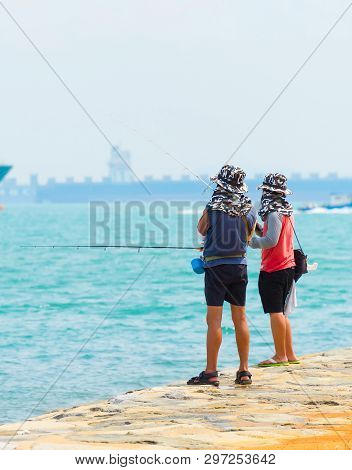 Fishermen Fishing From The Embankment Of Singapore Harbor. Commercial Cargo Ships In The Background