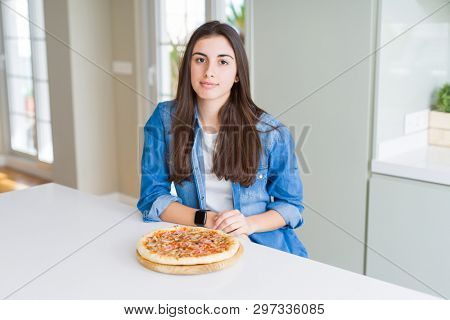 Beautiful young woman eating homemade tasty pizza at the kitchen with serious expression on face. Simple and natural looking at the camera.
