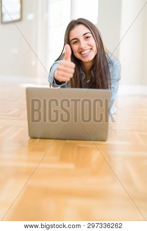 Beautiful young woman laying on the floor using laptop happy with big smile doing ok sign, thumb up with fingers, excellent sign