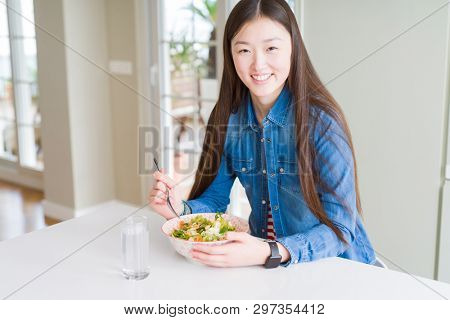 Beautiful Asian woman eating healthy pasta salad with a happy face standing and smiling with a confident smile showing teeth