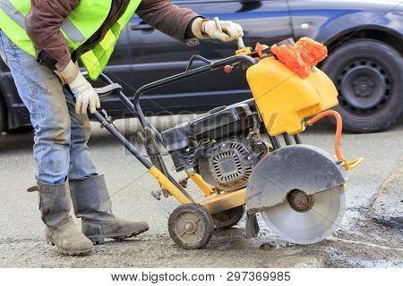 A Worker In A Green Reflective Vest Cuts A Piece Of Bad Asphalt With A Gasoline Cutter On The Carria